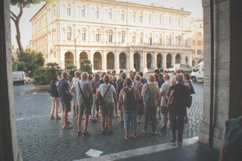 From inside a shaded arcade doorway, backs to camera, a tour group gathers on grey basalt cobbles of a broad piazza in central Rome; late morning sun rakes the Palazzo facade, a dropped paper map lies near a kerb.