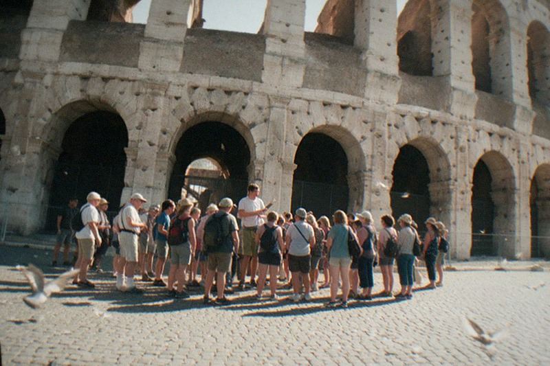 From a low angle near the arches on a hazy summer afternoon in Rome, a walking tour crowd circles its guide beneath the Colosseum's travertine arcades, pigeon feathers drifting across sun-bleached sampietrini cobbles.