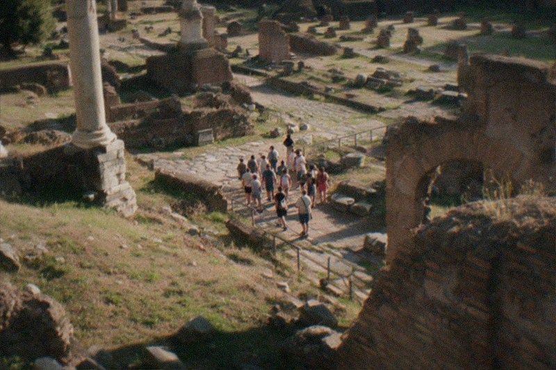 High on an overlook above the Roman Forum in Rome, Italy, an ancient-Rome walking tour stands as tiny distant figures with their backs turned, broken columns and cracked brick ruins below, low golden afternoon sun raking across the dusty stone.