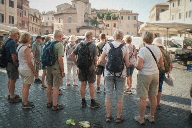 A two-hour free walking tour of Rome pauses at Campo de' Fiori in Italy on a hazy mid-morning; the group appears only as small distant backs on worn cobbles, market stalls blurred at the frame edge, a fallen lettuce leaf by the drain.
