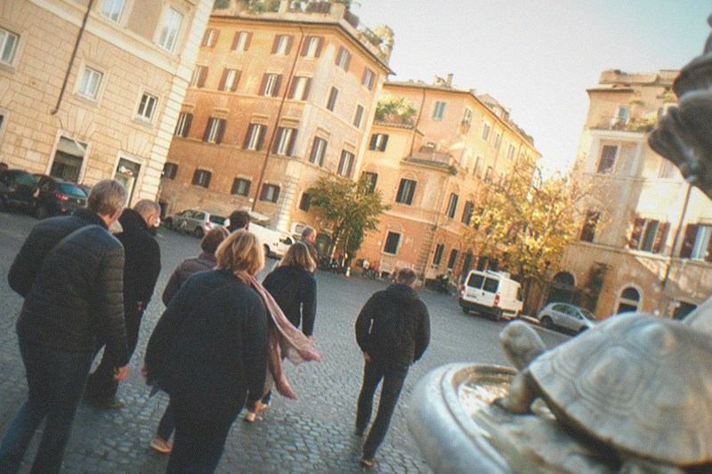 Seen from behind, a small tour group follows its guide across the worn sampietrini cobbles of Piazza Mattei in Rome, Italy, the turtle fountain blurred at the frame's edge, autumn afternoon light raking the ochre Renaissance facades, one walker's scarf trailing loose.