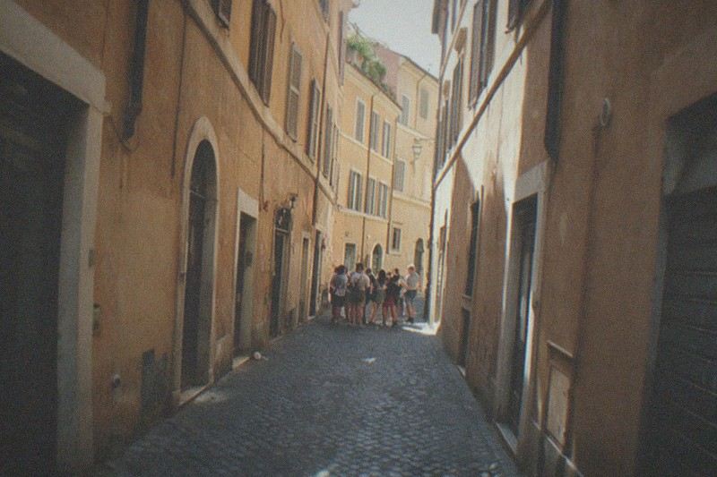 Midday summer light slants across a narrow cobbled lane of ochre Roman render near Campo de' Fiori in Rome, Italy; a small lunch group and their guide stand far off, seen only from behind, backs small against the worn sampietrini paving and shuttered facades, a stray paper cup by a doorway.