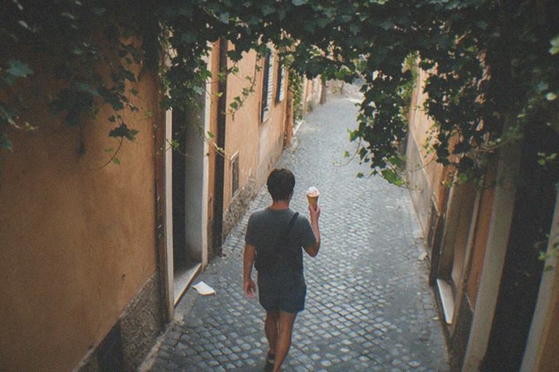 Overhead angle down a narrow cobbled Trastevere lane in Rome, Italy, ochre rendered walls and ivy overhead, a small distant figure seen from behind carrying a paper gelato cone, soft midday shade, a dropped napkin by a doorway far out of focus.