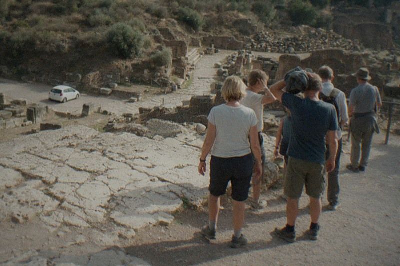 High on an overlook above the Roman Forum in Rome, Italy, a small walking group seen only from behind pauses on dusty gravel beside cracked travertine ruins; hazy afternoon light, one man wiping sweat with a folded jacket.