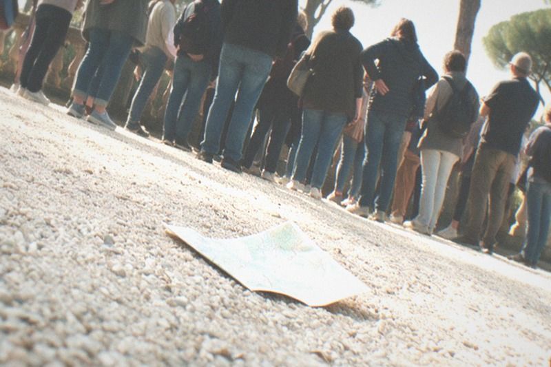 From a low angle on gravel, a tour group stands backs to the camera beneath the tall stone pines of Villa Borghese in Rome, Italy, soft spring haze over the umbrella pines, a classical balustrade filling the wide frame, a dropped paper map resting on the pale gravel.