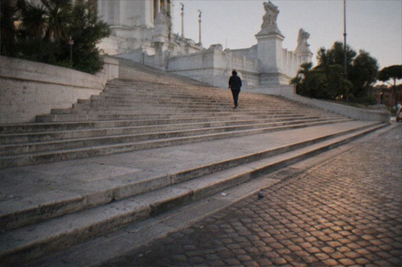 Dusk on the worn travertine steps of Piazza Venezia in Rome, Italy, a lone figure small and distant with their back turned, walking up toward the Vittoriano's pale marble terraces, lamplight warming the cobblestones, a scattered pigeon feather near the curb.