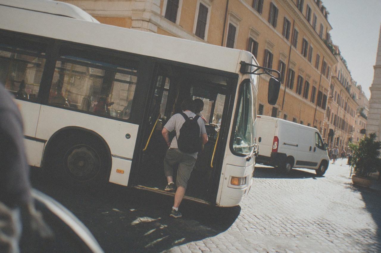 A city bus stopped at a sunny street corner in central Rome with baroque buildings behind it