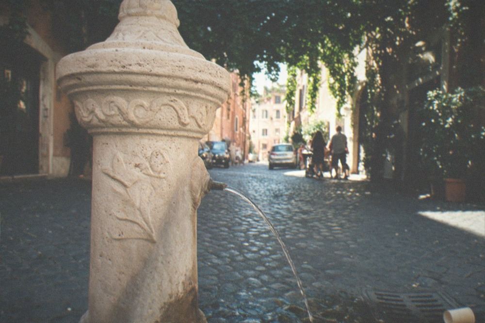 A stone fountain spouting drinking water beside a shaded Roman street