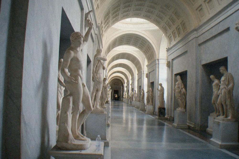 A long corridor of the Vatican Museums lined with marble statues and arched ceilings, filled with visitors looking at the exhibits