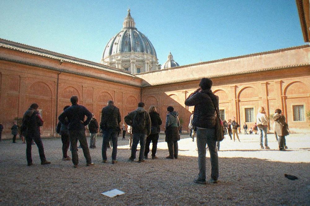 Visitors sitting on benches in a courtyard of the Vatican Museums with the dome of St. Peter's visible in the background under a clear blue sky