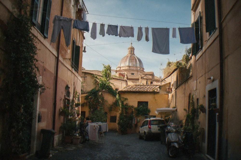 A narrow Roman alley with laundry hanging overhead and a distant church dome