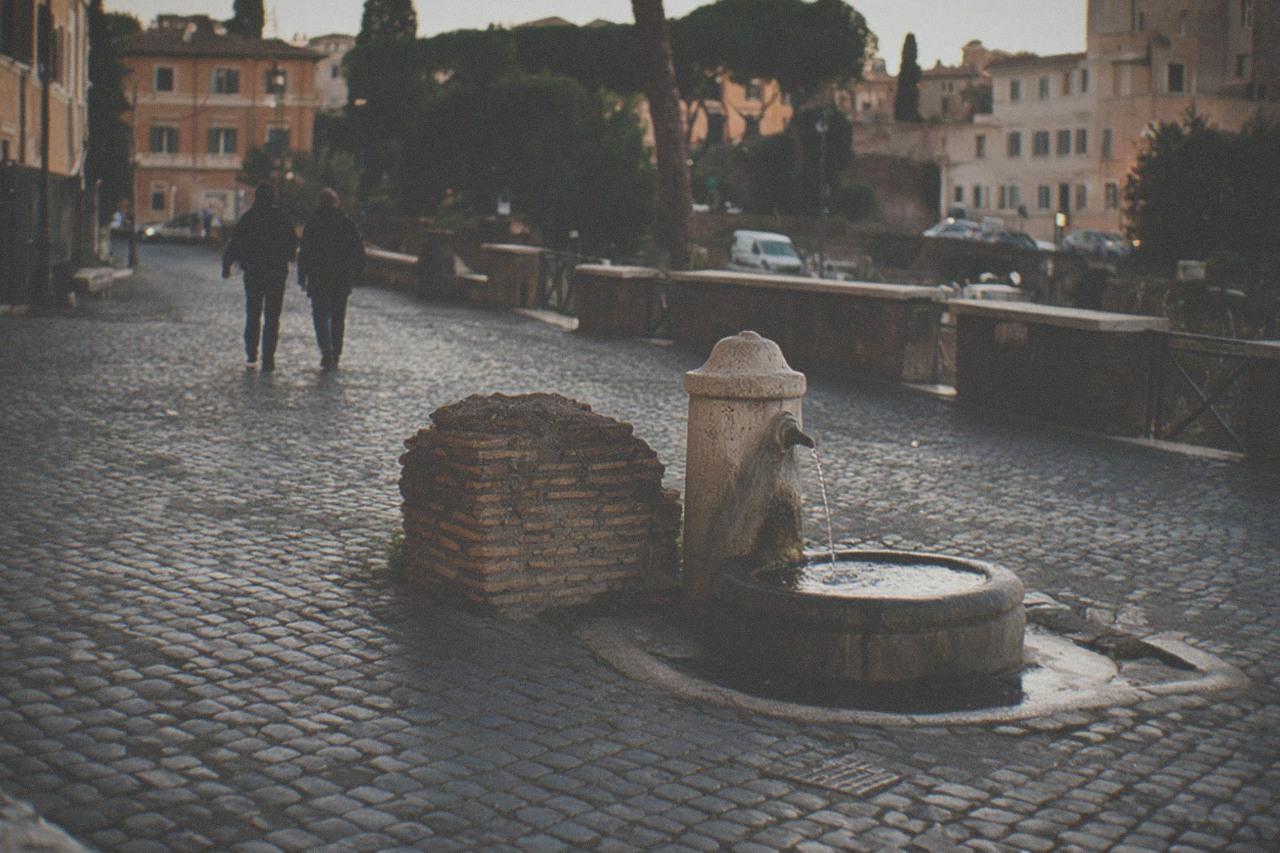 Cobblestone street in Rome's historic center with ancient ruins and a fountain under evening light.