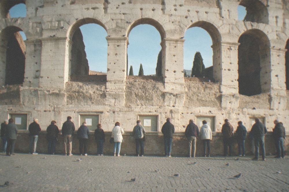 The Colosseum exterior with a line of visitors near the ticket office under a blue morning sky.