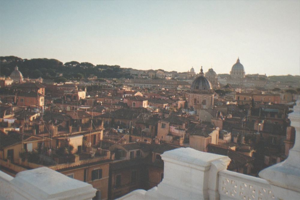 A wide view over Roman rooftops at sunset with a dome in the distance