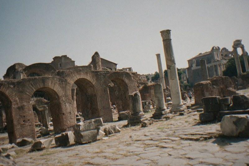 Weathered brick arches and broken marble columns of the Roman Forum under a hazy afternoon sky, dry grass pushing up through the cracked paving.