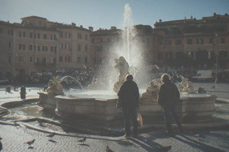 A baroque fountain with its stone tritons in Piazza Navona, late morning sun on the wet basin, pigeons scattering across the sampietrini cobbles below ochre Roman palazzi.