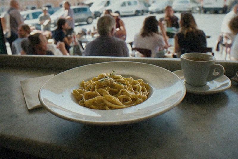 Fresh pasta and a small espresso cup on a scuffed marble counter in a plain Roman lunch spot, the busy room and the piazza outside falling away into blur.