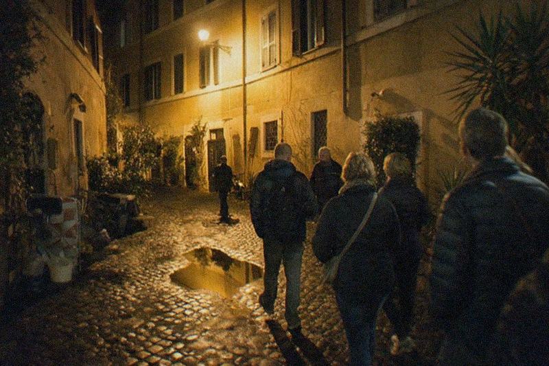 A narrow Trastevere lane after dark, wet sampietrini cobbles catching amber lamplight below shuttered ochre walls, a puddle mirroring the single street lamp.