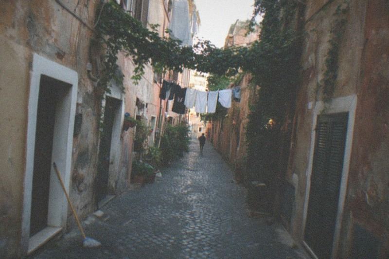 Ivy and washing lines cross above a cobbled Trastevere alley in Rome, ochre and rust plaster peeling from the old walls, one figure walking away down the lane in early evening light.
