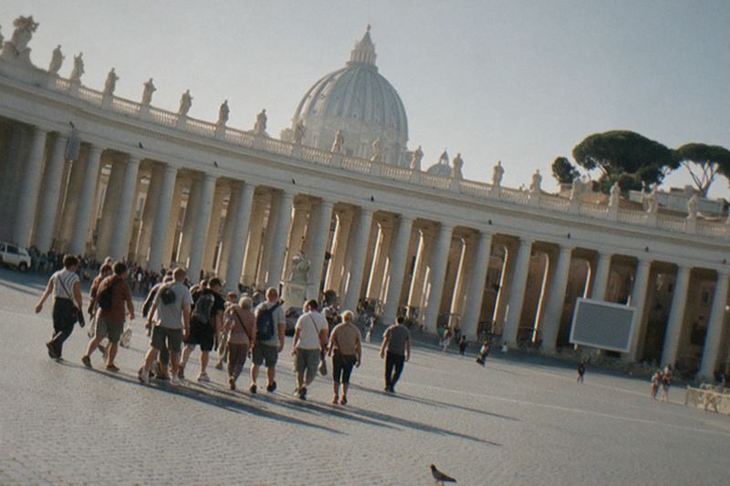 Bernini's curved colonnade and the pale dome of St Peter's above the wide cobbled square in flat early morning light, stone pines beyond and a pigeon crossing the empty expanse.