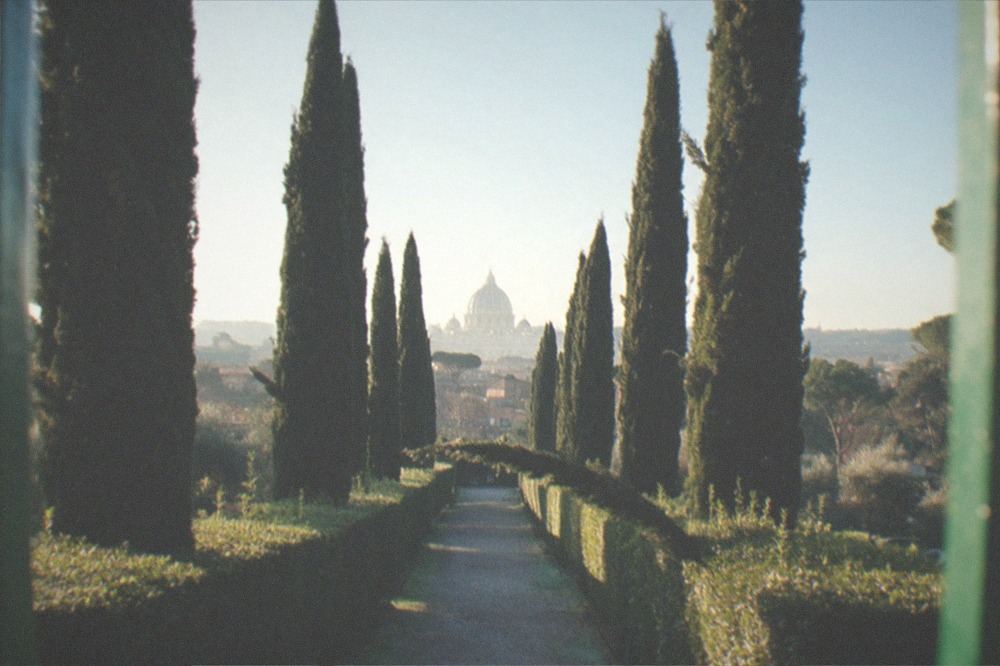 The Aventine keyhole seen through the green gate of the Priory of the Knights of Malta, with a perfectly framed view of St Peter's dome at the end of the hedge-lined path.
