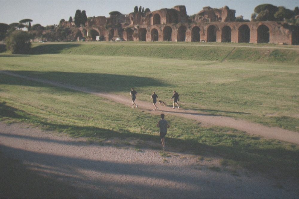 The Circus Maximus at sunset with the lawn stretching toward the Palatine Hill ruins, a few joggers and dog walkers crossing the open space.