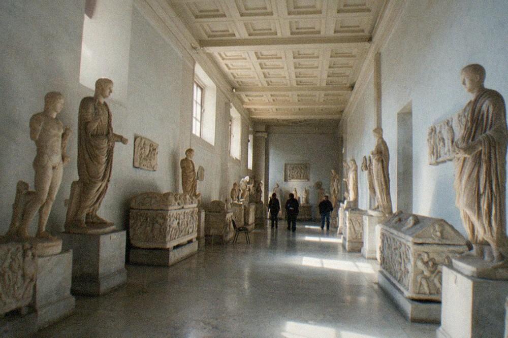 Sunlit gallery hall with ancient Roman statues and stone sarcophagi along both walls