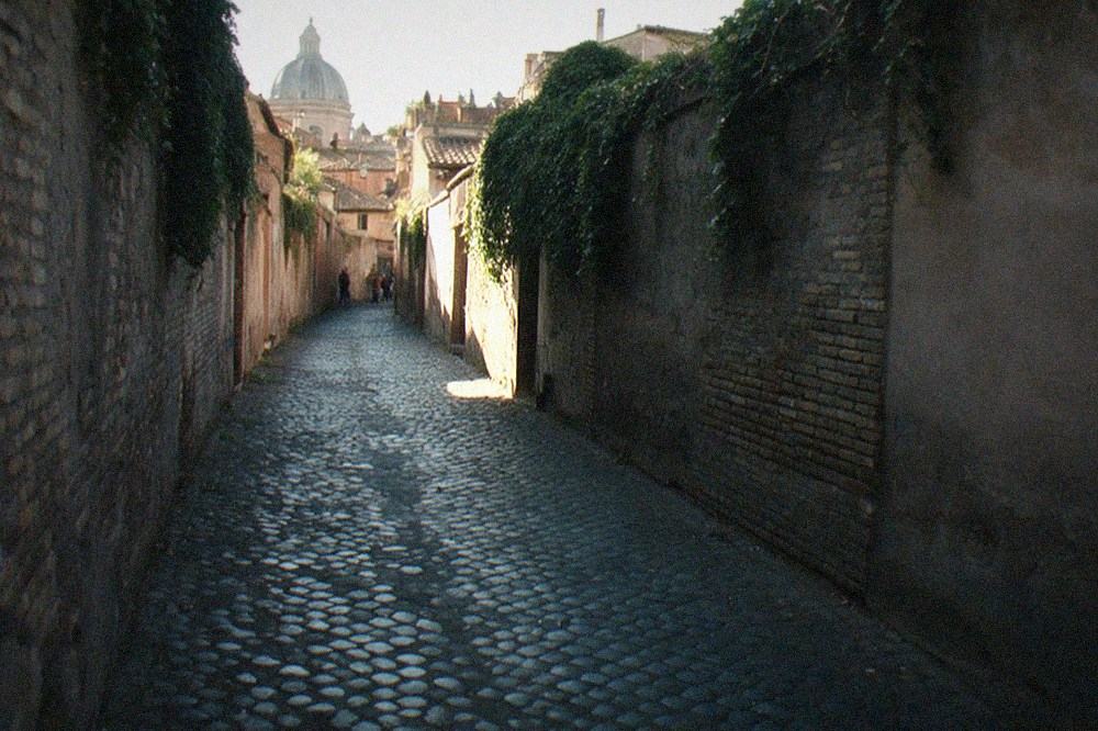 A quiet Rome alley with ivy-covered walls and a distant glimpse of a cupola.