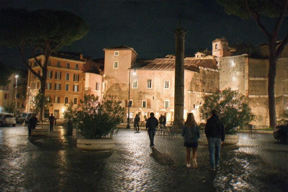 A brightly lit Rome piazza at night with the silhouette of an ancient column and people walking.