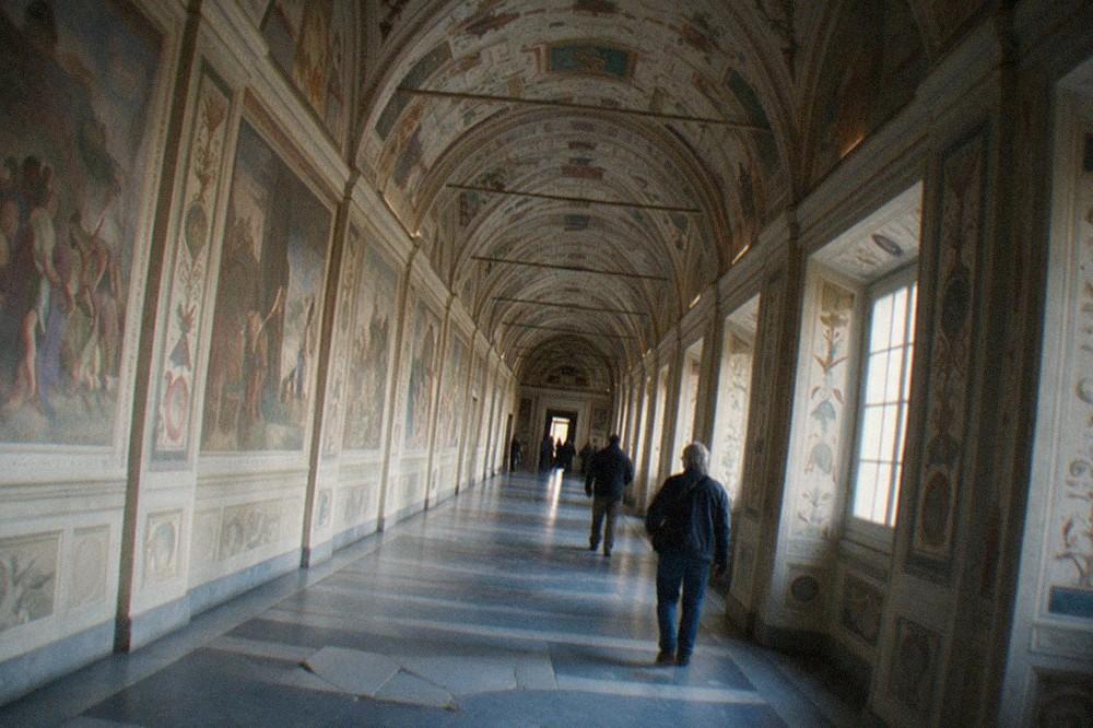 A long Vatican Museums gallery with frescoed walls and a painted ceiling leading toward distant doorways