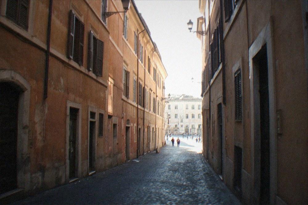 A narrow cobbled street in central Rome lined by ochre buildings leading toward a piazza