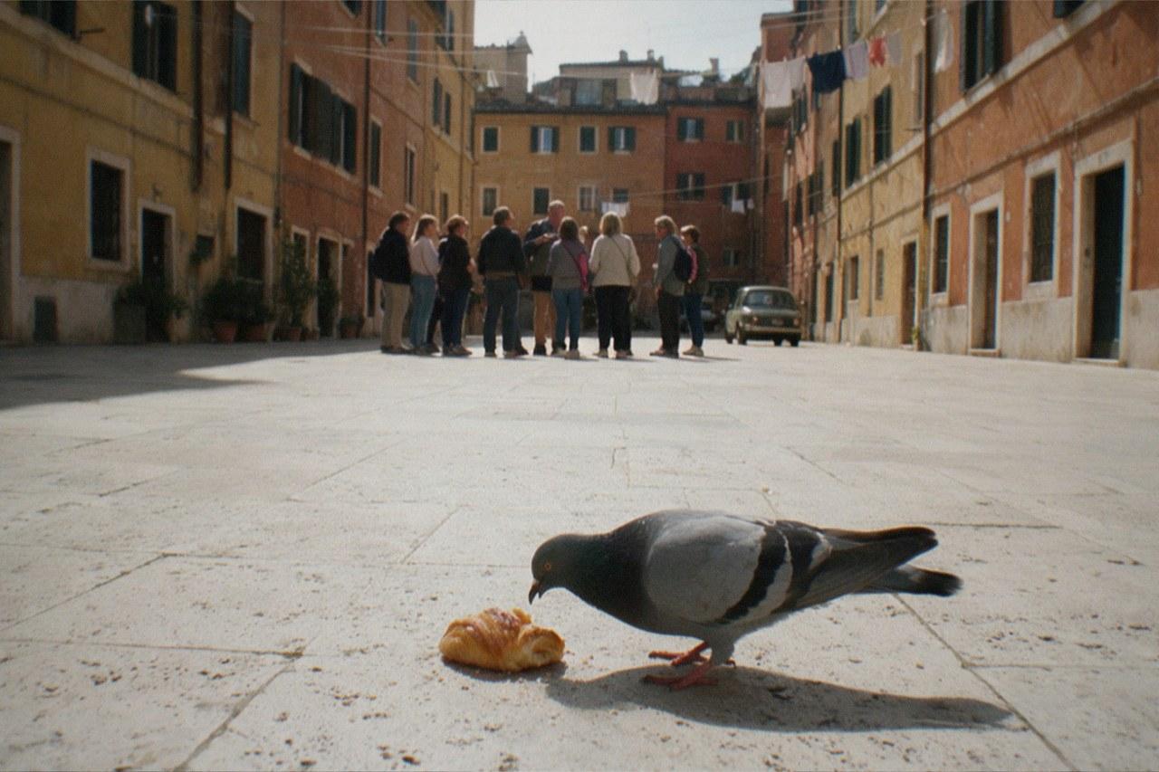 A small walking tour stands far down a travertine paved square in Rome, backs turned around their guide on a bright spring morning, ochre and terracotta facades and strung laundry cords filling the frame behind them.